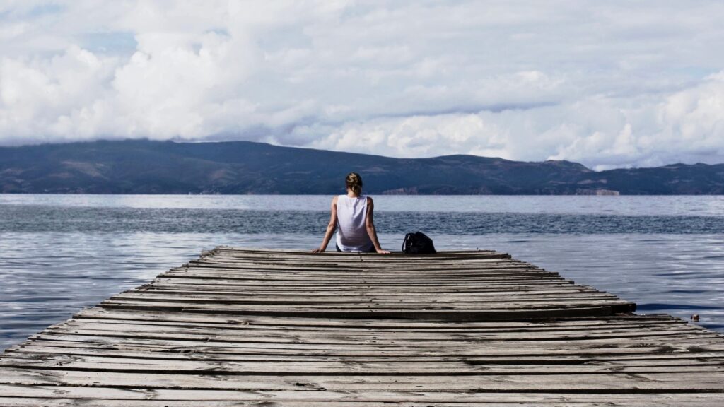 A woman sitting on the edge of a wooden plank infront of the ocean looking over the other island.